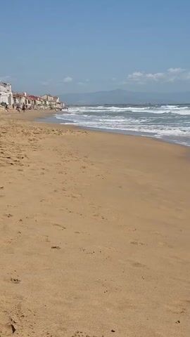 The video shows a bright sunny beach with light tan sand and waves coming in.  A line of buildings can be seen in the distance to the left along the beach. Some people are walking along the beach in the distance. Beyond the ocean, some mountains are faintly visible in the distance against a blue sky. At one point a child's voice can be heard saying "yeah yeah yeah yeah."