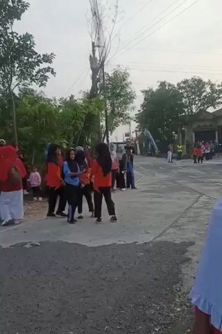 A group of people, mostly women and children, are gathered outdoors on a paved road. Some individuals are wearing red and white outfits, and others are in orange shirts and black pants. A few people are holding what appear to be musical instruments, possibly guitars or similar string instruments. The atmosphere seems casual and social, with people interacting and observing each other. In the background, there are trees, a building with a tiled roof, and utility poles with wires. The lighting suggests it is either early morning or late afternoon.