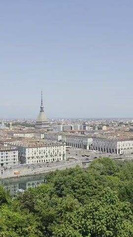A daytime shot shows a cityscape under a clear, pale blue sky. The foreground is occupied by lush green trees. Beyond the trees, a river or body of water reflects the sky and nearby buildings. The city's buildings, predominantly white or light-colored with red-tiled roofs, stretch into the distance. A prominent architectural feature is a tall, slender tower with a spire on top, centrally located in the view. The tower stands out against the surrounding buildings due to its height and distinctive design. In the far distance, faint mountain ranges are visible along the horizon. The camera pans slightly to the left, revealing more of the city's layout and the buildings along the riverfront.
