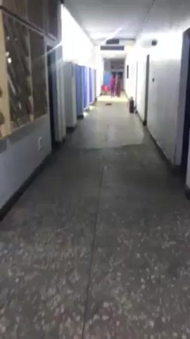The video shows a long, dimly lit hallway with white walls and a gray, speckled floor. On the left side of the hallway, there are several cubicles with blue curtains. On the right side, there are closed doors. Further down the hallway, two people in dark clothing are walking away from the camera. At the end of the hallway, there is some red furniture and a sign overhead. A fire extinguisher is also visible on the right side of the hallway. The camera moves slightly, providing a view from the end of the hallway.