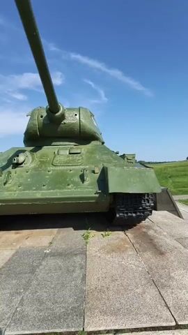 A low-angle, close-up shot of a green Soviet T-34 tank. The tank is on a stone base with some weeds growing between the stones. The sky above is blue with a few wispy white clouds. The tank's long gun barrel points upwards and to the left. The camera pans slightly to the right.