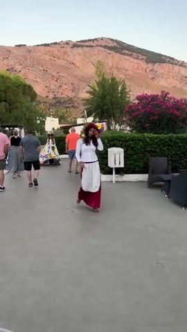 In the video, a woman is walking towards the camera. She is wearing a white long-sleeved shirt, a red skirt, and a white apron. She has a large floral headdress on her head. In the background, another woman in a colorful dress is walking away from the camera, followed by a man in an orange shirt and shorts. Further in the background, a group of people are walking to the left. The scene appears to be outdoors, with a paved walkway and a hedge lining the right side. A rocky mountain is visible in the distance behind the people and foliage. The lighting suggests it is late afternoon or early evening.