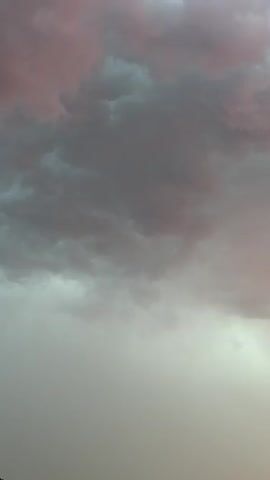 The video shows a close-up, vertical view of a cloudy sky. The clouds are dark gray and appear heavy, with some hints of pink and purple on the edges, possibly indicating a sunset or sunrise. The bottom portion of the video is a lighter shade of gray or beige, suggesting the sky transitioning to a clearer area or the horizon. There are no people, objects, or discernable actions visible in the frame.