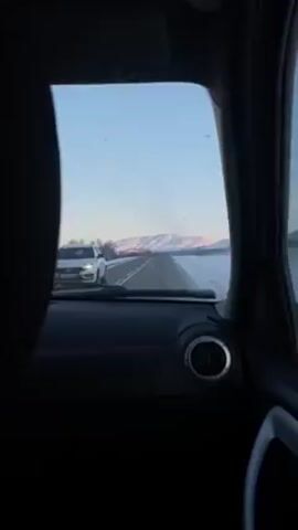 The view from inside a car shows a straight paved road ahead, flanked by snow-covered ground. In the distance, snow-capped mountains are visible under a pale blue sky. The car's dashboard is visible, with circular air vents and a wiper blade resting on the windshield. The overall lighting suggests it is early morning or late afternoon.
