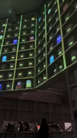 The video shows a large room with high ceilings and multiple shelves reaching towards the ceiling. The shelves are full of boxes or containers, and a projector displays images of Pac-Man ghosts and simple ovals onto the boxes. People are visible below the shelves, near what appears to be a counter, suggesting a commercial or entertainment space. The overall lighting is dim with the projection lighting illuminating the shelves.