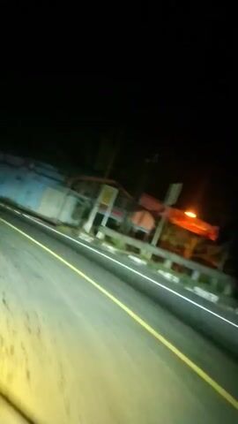 This is a blurry, shaky video shot from the perspective of someone in a vehicle at night. A road with a yellow line down the middle is visible. On the right side of the road, there is a long, low building with sections of blue and white. The building appears to have some colorful elements, possibly graffiti or murals, in shades of purple and pink. There are also some orange elements visible on the building, and some red accents on railings or structural components. Beyond the building, there is a dark, wooded area. The headlights of the vehicle illuminate the road ahead. The video is dark and low quality, with motion blur.