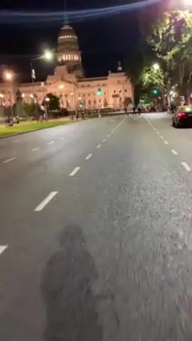 The video shows a view from a moving vehicle on a road at night. The road has white lane markings. A large, lit building with a dome is visible in the background. Pedestrians are present near the building. A red car is on the road. Streetlights illuminate the scene. Trees are visible along the side of the road.