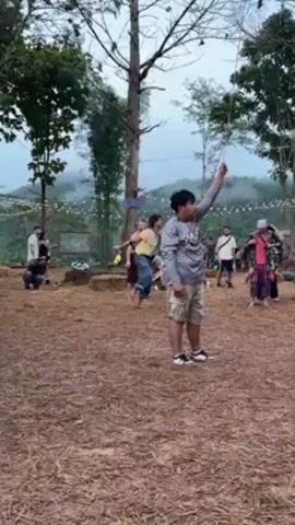 The video shows an outdoor gathering of people on an area covered in pine straw, with trees and what looks like a stage or setup in the background. A boy in a grey shirt and khaki shorts is standing near the center, holding a string connected to a birdhouse suspended in the air. A man with curly hair walks in front of the camera, briefly obscuring the view. Several people are standing and mingling in the background, some taking photos. The environment suggests a casual, festive atmosphere. There are lights strung between the trees. The terrain behind the crowd slopes up to a mountain covered with fog.