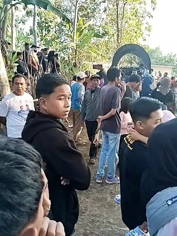 The video shows a gathering of people outdoors, possibly in a rural or semi-rural setting. There are trees and vegetation in the background. In the foreground, a group of people are standing and interacting. One person in the foreground is wearing a black hoodie and has their arms crossed. Further back, a man in a grey jacket is holding up a phone, seemingly recording or taking a picture. Several people are wearing headscarves, suggesting a community or religious event. There are also some objects on the ground, including buckets and what appears to be a water source being filled from a larger container. The overall atmosphere seems informal and social.