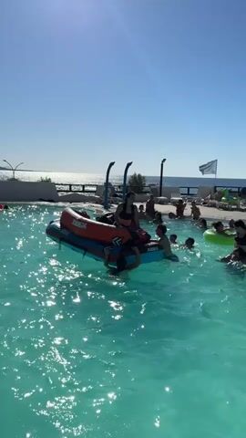 In a swimming pool with clear blue water and bright sunlight, several people are enjoying a hot day. In the foreground, an inflatable raft, blue with orange accents, is in the center of the pool. A young woman, wearing a black bathing suit, is seated on the raft, appearing to steer it. Another young person is partially submerged in the water, reaching towards the raft. Around the raft, other individuals are swimming and playing. In the background, more people are visible in the pool, some on inflatable rings. Beyond the pool, lounge chairs and a white railing can be seen. The sea and a bright sky form the backdrop, with the sun glinting off the water. A large, colorful beach ball is in the air, heading towards the raft. The overall atmosphere is one of leisure and fun.