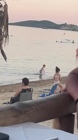 The media file depicts a beach scene at dusk in Nea Peramos, Greece. In the foreground, a group of people are relaxing on the sand. To the left, a person sits in a camping chair, while a child in a pink bikini plays near a blue beach chair. Further back, several people are enjoying the water, with some swimming and others standing near the shore. In the distance, a hazy silhouette of land with a structure on it is visible across the calm sea. The sky displays soft hues of pink and orange, indicative of sunset. The overall atmosphere appears relaxed and leisurely, with families and individuals enjoying a pleasant evening at the beach. There is no visible text in the media file.