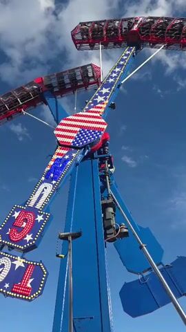 The video shows a thrill ride at an amusement park. The ride is predominantly blue and red, with American flag motifs on its arms and a cage-like passenger car decorated in red. The ride is in motion, swinging and rotating against a bright blue sky with scattered white clouds. In the background, other amusement park attractions, including a green and purple ride, can be seen, along with some buildings. People are visible within the passenger car of the ride, appearing to be enjoying the experience. The camera is positioned below the ride, looking upwards at its ascent. The video captures the dynamic movement and scale of the amusement park attraction.