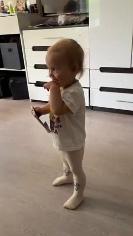A young child, appearing to be a toddler, stands in a room, holding a smartphone. The child is wearing a white t-shirt with colorful text that is difficult to read, and light-colored leggings with a patterned design on the lower legs. The child's hair is blonde. The child's right hand is raised to their face, near their mouth, and they appear to be looking down at the phone. The room has light-colored wooden flooring and white furniture, including a dresser with multiple drawers and a tall white cabinet. There are also some dark storage bins visible. The overall impression is that of a domestic setting. The child's facial expression is a bit ambiguous, but their posture and hand gesture suggest a moment of absorption or perhaps shyness. There is no indication of the time of day or weather conditions. The location context provided is Kumyonki, Russia.