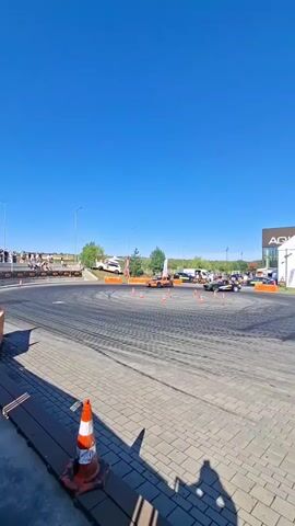 Two cars are drifting on a circular asphalt track. The track is surrounded by a fence and cones. In the background, there are spectators and buildings. The sky is clear and blue.
