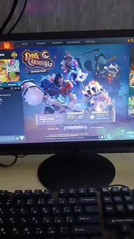 A computer monitor displays the game Dota 2, with the "Dark Carnival" event banner visible. The game interface shows character portraits and event details. In front of the monitor sits a black keyboard with a numeric keypad. To the right of the keyboard, on the desk, are an energy drink can, a bottle of water, and a white vape device. The scene appears to be a gaming setup.