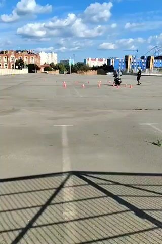 The video shows a motorcycle rider practicing on a large, flat asphalt lot. The rider is wearing a helmet and riding gear and is maneuvering through a course marked by orange traffic cones. In the background, there are several buildings, including some older brick structures and more modern multi-story buildings. A blue construction crane is visible in the far distance. The sky is blue with scattered white clouds, suggesting a pleasant, possibly daytime, weather condition. The overall scene appears to be a training area or an empty lot used for practice. There is no visible text in the video.