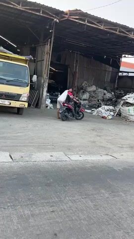 A truck is parked on the left side of the image. A motorcycle is in the middle, with two people on it. One person is driving, and the other is holding a white bag. The motorcycle is facing away from the camera. In the background, there is a building with an open roof, and piles of garbage and debris. The ground is paved.