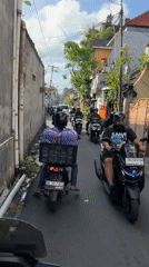 Motorcyclists navigate narrow Bali street, one carrying purple load