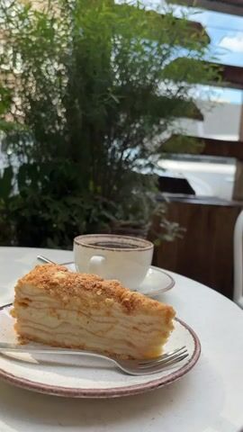 A slice of Napoleon cake and a cup of black coffee are on a white table outdoors. The cake is layered with cream and topped with crumbs. The coffee is in a speckled white cup with a saucer and a spoon. Behind the table, a large potted plant with green leaves is in a wicker basket. In the background, wooden beams and a white chair are visible. Beyond that, through an opening, a white car and a building are seen. The scene appears to be a cafe or restaurant with an outdoor seating area. The lighting suggests it is daytime.