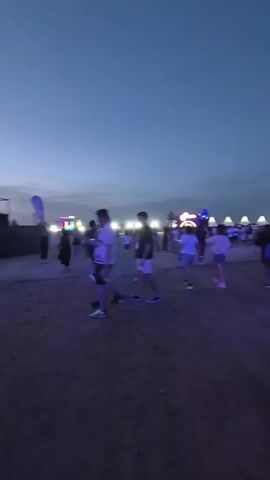 The video shows an outdoor event at dusk or night. In the foreground, a person wearing a red, white, and blue tank top and denim shorts is walking away from the camera. Behind them, several people are visible, including a group of four who appear to be interacting. Further in the background, there are tents with red canopies, stage lighting, and other festival-like structures. The sky is a deep blue, transitioning from daylight to night. Purple and blue lights illuminate parts of the scene. The ground is dirt or gravel. The overall atmosphere appears to be that of a concert or festival.