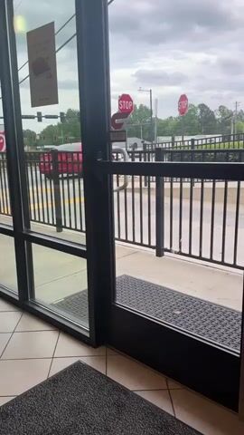 The camera, from an interior point of view, presents a view through the partially open glass door of a Chick-fil-A restaurant. A gray patterned rug is visible in the foreground. The door frame is black.  Outside, a black metal fence lines the sidewalk. Beyond the fence, a street with a red car is visible. In the distance, two stop signs are clearly visible. Traffic lights can be seen with red lights illuminated. The sky is overcast and gray. A poster can be seen attached to the glass of the door on the left. The floor inside the restaurant is tiled with a light beige and white pattern. The overall lighting is diffused.  Sounds of voices can be heard in the background.