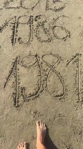 A person's feet are visible in the lower part of the frame, standing on a sandy beach. In the sand, several years are written: 2015, 1986, 1981, 2017, 2022, and 2014. The video pans upwards, showing more of the sand and the written years.