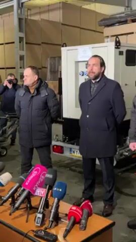 Here's a detailed description of the video:

Two men stand in front of a group of microphones, likely preparing for a press conference. The man on the left is wearing a dark puffer jacket with a brown turtleneck sweater underneath. The man on the right is wearing a dark overcoat. Both men have their hands clasped in front of them. 

Behind them is a white generator on a trailer with a Polish license plate. The background shows large stacks of what appears to be wood or plywood. The setting looks like a warehouse. 

In front of the men, on a table, there are several microphones from various news organizations, including those from Kyiv24, World, and TRT World. Another man stands to the left, partially visible, with a camera.
