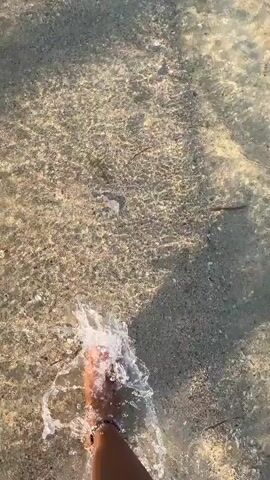 The video shows a first-person perspective of someone walking in shallow, clear water over a sandy bottom. The water is rippling and shows reflections of light. Small splashes are created as the person's feet move through the water. The person appears to be wearing dark-colored shoes or is barefoot, with toes visible. The environment is a natural body of water, likely a beach or a shallow riverbed. The footage is shaky, indicating movement.