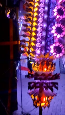 The video displays a nighttime scene featuring a decorated structure or vehicle adorned with numerous colorful lights. The lights are arranged in rows and patterns, with prominent purple, yellow, and pink hues dominating the display. Some lights are circular or star-shaped, while others form intricate geometric patterns. The structure itself appears to be elongated and possibly curved, with a layered design. Some sections have a feathery or fringed appearance, possibly part of the decoration. The overall impression is one of a festive or celebratory display.