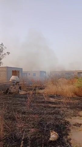 The video shows a field that has been recently burned, with patches of black ash and partially charred vegetation. In the background, there are several unfinished brick buildings with visible window openings. Smoke billows from behind the buildings, indicating a recent or ongoing fire. An old cement mixer sits to the left of the frame. The sky is overcast and hazy, potentially due to the smoke. The overall scene suggests an aftermath of a fire in a construction or rural area.