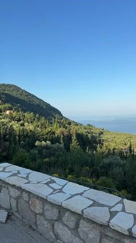 The video shows a panoramic view of a lush, green landscape leading to a calm sea under a clear blue sky. The foreground features a stone wall constructed with irregular, light-colored stones set in a dark mortar. The view extends over a forested hillside, predominantly green with interspersed taller, thin trees. Beyond the hillside, the sea stretches to the horizon, appearing flat and calm under the bright sky. The overall scene evokes a sense of tranquility and natural beauty.
