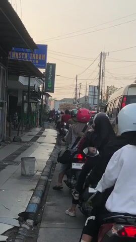 The video shows a street scene with several people on motorcycles. The motorcycles are lined up along the side of the road, suggesting they are waiting. The people are wearing helmets and some are carrying bags. There are buildings on the left side of the street, one of which has a sign that says "HASTA INDAH AIR." There are also power lines and a utility pole visible. The sky appears to be overcast or hazy. The overall impression is a busy street with a lot of motorcycle traffic.