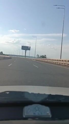 The video shows a view from inside a vehicle driving on a highway. The highway is a multi-lane road with a clear blue sky and scattered clouds overhead. The weather appears to be pleasant and sunny, suggesting it is daytime.

The primary subjects are the highway itself and the surrounding landscape. The road stretches out ahead, curving slightly to the left. On the right side of the highway, there is a guardrail and beyond that, a body of water, likely a river or a lake, with trees lining the far bank. On the left side of the highway, there are a series of lampposts and a large, empty billboard structure. The billboard is a prominent object, standing tall against the sky. The interior of the vehicle is visible in the foreground, showing the dashboard and windshield.

The activity is the act of driving on the highway. The perspective of the video indicates the camera is mounted or held within a moving vehicle. There are no people visible in the video.

Notable details include the clear weather and the presence of the empty billboard, which might suggest a location where advertisements are placed. The road markings and guardrails are typical of a highway. The scene evokes a sense of travel and open road. The time of day is clearly daytime due to the bright sunlight and visible shadows.

There is no visible text in the video. The location context provided is Yelyzavetivka, Ukraine, which helps to place the scene geographically.