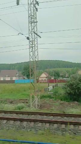 The video shows a view from a moving train traveling through a rural area. The foreground features a set of train tracks, while the middle ground displays a grassy field with several houses and fences. In the background, a densely forested hill or mountain range is visible under a cloudy sky. The houses appear to be of various colors and sizes, with one prominent blue house and others in lighter colors. The overall scene depicts a peaceful, suburban or village landscape passing by.