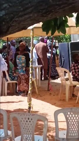 The video captures a scene under a tent, likely an outdoor event, possibly a celebration, where several people are gathered. A woman stands out, carrying a child wrapped in a blue and white cloth carrier. Various individuals are milling about, some dressed in traditional African attire, while others are in more contemporary clothing. The environment appears to be a sandy or dirt surface, with rows of white plastic chairs set up, suggesting a seated gathering. Speakers are visible, indicating music or announcements are part of the event. The overall atmosphere seems lively, with conversations and movement among attendees.