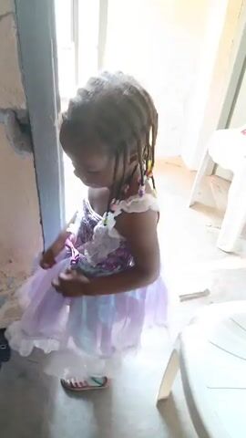 The video shows a young child dressed in a purple and white dress. The child has dreadlocks with colorful beads. The child is standing in what appears to be a doorway. A white plastic chair is visible behind the child. The child touches the wall briefly. The child looks around. Music is playing in the background.