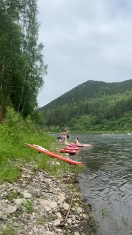 The video shows a group of people on a rocky shore next to a wide, calm river. Several red inflatable paddleboards are lined up on the grassy bank. In the water, a few individuals are on paddleboards, and one person appears to be sitting on the edge of a paddleboard. The background features a densely forested hillside under a cloudy sky. The scene suggests a recreational activity like paddleboarding or kayaking in a natural, outdoor setting.