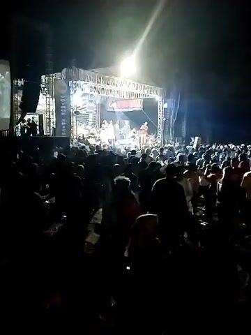 A live music concert is taking place outdoors at night. A band is performing on a raised stage, illuminated by bright spotlights and colored stage lighting. The stage is adorned with banners and equipment, including speakers and musical instruments. A large crowd of people is gathered in front of the stage, facing the performers. Many in the crowd appear to be dancing and enjoying the music, with some raising their hands in the air. The overall atmosphere suggests a lively and energetic event. The environment is dark, typical of an outdoor night performance, with the stage lights being the primary source of illumination in the immediate vicinity.