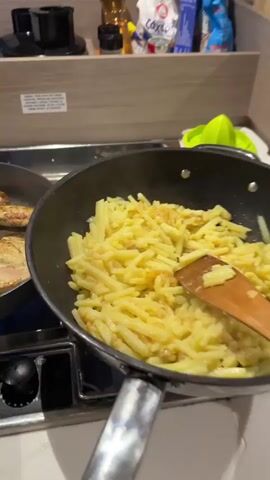 A first-person view shows a kitchen counter with food being cooked. In a pan on the left, several pieces of browned chicken are cooking. In a pan on the right, some yellowish pasta is being stirred with a wooden spoon. Behind the pans, a black coffee maker sits on the counter. The camera pans right, revealing a reflective oven door, which shows reflections of the kitchen, including a wooden cutting board and some knives. The camera continues to pan right, showing a window that looks out onto a dark area, possibly outside, with some lights visible in the distance. The window also reflects the interior of the kitchen, showing some items on a table and a person in the background.