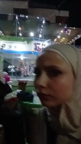 The video shows a young woman wearing a beige hijab in an outdoor setting at night. At the beginning of the video, she is seated and looks at the camera with a slight smile. The camera pans to show the surrounding area, including string lights, a building facade, and other people. In the later part of the video, the woman is seated at a table, examining something in her hands. There's a stone wall and some greenery in the background.