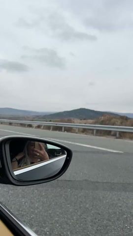 Here is a concise description of the video:

The video shows the side mirror of a moving car, reflecting a woman with blonde hair who is holding up a phone and taking a picture of herself. The car is on a highway, with a white truck passing on the left. The highway has a center line, and a metal guardrail separates the road from the landscape. In the distance, there are rolling hills under a cloudy sky.