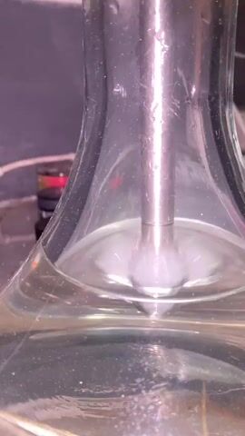 This is a close-up, still shot of a glass container filled with a clear liquid. A dense cloud of white vapor or smoke is bubbling up from the bottom of the container, creating frothy effervescence as it rises. The vapor is thick and opaque, obscuring the view of the lower portion of the glass. Small bubbles are also visible in the liquid itself. The lighting creates a glare on the glass, and there are droplets of moisture on the exterior surface of the container. In the blurred background, what appears to be a dark bottle with colorful bands and some wires are faintly visible. The overall impression is of a chemical reaction or a process involving the vaporization of a substance within the liquid. There is no discernible text in the image. The context provided suggests this is from Ipatovo, Russia.