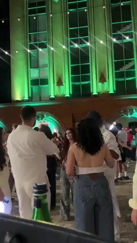 The video shows an outdoor gathering of people at night. The scene is illuminated by artificial lights, which change color from green to blue, casting a vibrant glow on the building in the background. The building appears to be a large, multi-story structure with a tiled facade and arched doorways. String lights are visible overhead. In the foreground, a group of people are socializing and dancing. Some individuals are wearing white clothing, while others are in casual attire. In the lower part of the frame, a bottle with a green cap is partially visible. The overall atmosphere suggests a lively party or event.