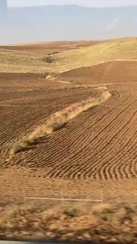 The video shows a view of cultivated fields from a moving vehicle, likely a train or car, as evidenced by the blurred vertical lines of window frames in the foreground. The landscape consists of rolling hills with patches of brown, tilled earth and some areas of lighter colored vegetation. The tilled fields exhibit distinct patterns of rows and furrows, suggesting recent agricultural activity. A thin wire fence is visible in the lower part of the frame, running horizontally across the scene. The sky is pale and hazy, and the overall impression is of an agricultural expanse under natural light.