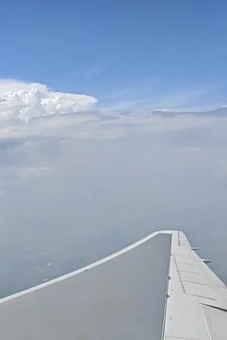 The video shows a view from an airplane window, looking out at the sky. The sky is blue and partly cloudy, with large white clouds dominating the scene. The wing of the airplane is visible in the foreground, with its white surface and flap mechanisms clearly seen. Below the clouds, a hazy landscape can be perceived, suggesting the airplane is at a significant altitude. The clouds appear to be cumulonimbus, with some possibly developing into thunderstorms. The overall impression is of a vast expanse of sky viewed from above the clouds.