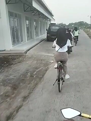 A group of four individuals ride bicycles along a narrow concrete path beside a long, low, grey building. The first cyclist, viewed from behind, wears a white shirt, grey pants, and a dark headscarf. The second cyclist, also viewed from behind, wears a white and black shirt with dark pants. The third cyclist wears a light green shirt with dark pants. The fourth cyclist wears a blue top and white shorts. To the side of the path, near the building, a small black truck is parked. A man stands near the truck. Beyond the path, there's a body of water and flat terrain stretching to the horizon under an overcast sky.