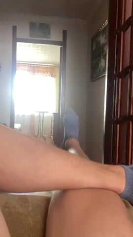 The video shows a close-up view of a person's legs and feet, viewed from a first-person perspective. The person appears to be lying down, possibly on a couch, as indicated by the textured fabric visible in the foreground. Both legs are extended upwards, with the feet wearing grey socks. One leg is closer to the camera and takes up a significant portion of the frame, while the other leg is slightly further away.

The background suggests an indoor environment. A doorway or archway is visible in the mid-ground, leading to another room or area. Through this opening, a window with sheer curtains is illuminated by bright sunlight, creating a washed-out effect. A small table with what appears to be a tripod or stand is also visible in the illuminated space.

To the right of the frame, a wooden door with glass panels is partially visible. On the wall above this door, a framed picture or tapestry with a green landscape scene is hanging. The walls in the background appear to be a neutral color. The overall lighting is somewhat dim, except for the bright light coming from the window in the background.

The audio consists of a male voice speaking in Russian. The person's voice is calm and conversational. The speech appears to be describing the action in the video or a related topic. The spoken words include phrases like "Let's put it in" and "these are separate side dishes," suggesting a discussion about food or a meal, or perhaps about items being placed somewhere.