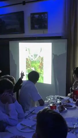 The video shows a projector screen displaying a series of images. The first image is of a young woman in a white top and a red and white patterned skirt, with a red bandana in her hair, standing in a grassy outdoor setting. She is looking to her left and her arms are outstretched. The second image is of a woman with blonde hair, wearing a white shirt and a beige vest, sitting at a desk with some cabinets and a blue bin behind her. She is looking to her right. The projector is in a dimly lit room. In the foreground, several people are seated at a table, facing the screen. The lighting in the room is predominantly blue. Some of the people are silhouetted. There are plates, glasses, and a pitcher of red liquid on the table.