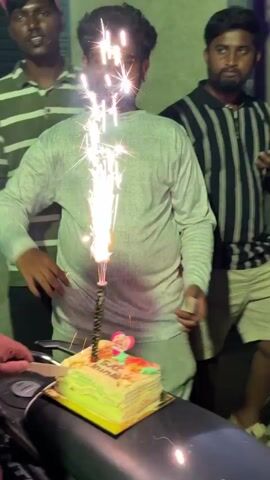 A group of South Asian men celebrate a birthday outdoors in the evening.  The video opens with three men in front of a motorcycle, on the seat of which sits a cake with a lit sparkler candle.  The man in the middle of the frame is wearing a white shirt and smiling, preparing to cut the cake.  The other men around him are singing, and another man in the background is taking a video.  As the camera pans and the video continues, the frame fills with five young men. The birthday cake is out of focus. The men stand on a street and clap.