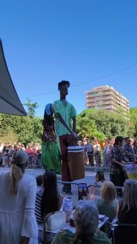 This video shows a sunny outdoor street scene with a parade or performance. Two giant puppets are the focus of the action, one dressed in a green patterned shirt and red pants, holding what appears to be a drum. The other puppet is adorned in a floral patterned top, a long green skirt, and a blue head covering. They are positioned in the street, with a crowd of onlookers gathered behind a metal barricade. Many people in the crowd are holding up their phones, presumably to record the event. The background reveals trees and a multi-story building, suggesting an urban or semi-urban environment. The sky is clear and blue. Several people are seated at tables in the foreground, some also observing the performance.